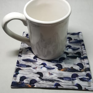 May include: A white ceramic mug with a handle sits on a gray and white fabric coaster with a pattern of black and blue loon birds.