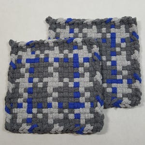 May include: Two woven potholders in a square shape. The potholders are gray, light gray, and blue, with a woven checkerboard pattern. The edges are finished with a braided border. The potholders are likely made of cotton or a similar material.