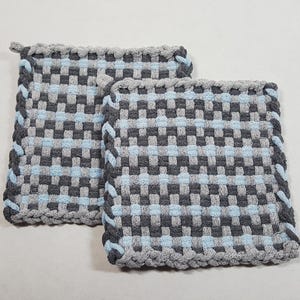 May include: Two square woven potholders. The potholders are gray, light blue, and dark gray with a braided gray border. The potholders are made of a woven material and are designed to protect surfaces from heat.