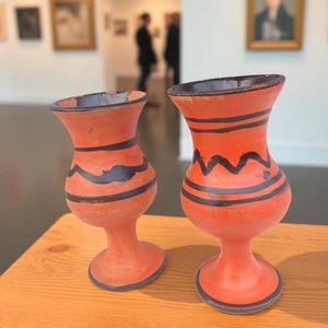 May include: Two terracotta vases with a vibrant orange hue and dark blue accents. Each vase features a unique design of horizontal and zigzag lines. The vases are displayed on a wooden surface, with a gallery background.