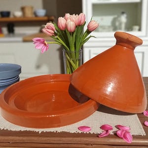 Può includere: Una pentola tagine aperta, marrone-arancio, con coperchio conico, esposta su una superficie di legno. La pentola è accanto a un vaso di tulipani rosa. Il tagine è un tradizionale recipiente da cucina nordafricano.