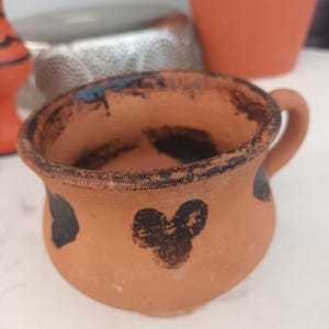 Puede incluir: Una pequeña taza de terracota hecha a mano con asa. La taza presenta un tono natural y terroso con detalles pintados en negro. El borde y el asa tienen un aspecto más oscuro, ligeramente quemado. La taza tiene una forma redondeada y un diseño simple y rústico.