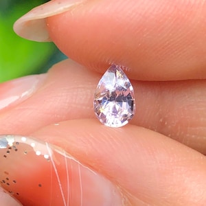 May include: A pear-shaped, faceted gemstone with a pale pink hue. The gem is held between fingers, showcasing its clarity and brilliance. The cut of the stone allows light to reflect, creating a sparkling effect. This gemstone is suitable for jewelry.