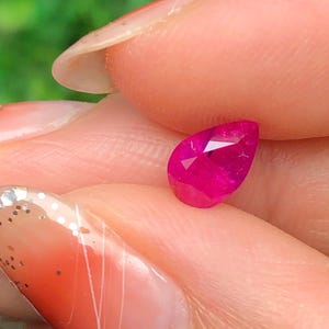 Peut inclure: Un rubis en forme de goutte d'eau, facetté. La gemme est d'une couleur rose-rouge vive et saturée, avec des facettes internes visibles. Le rubis est tenu entre les doigts, avec un arrière-plan vert flou.