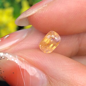 Puede incluir: Una pequeña gema facetada de forma ovalada con un cálido tono amarillo-naranja. La gema se sostiene entre los dedos, mostrando su superficie pulida y claridad interna. El fondo está borroso, resaltando el color y la forma de la gema.