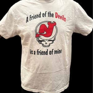 May include: White t-shirt with a crew neck featuring the text "A Friend of the Devils is a friend of mine." The design includes a red "N" logo with a devil's tail inside a circle, above a skull.