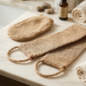 Puede incluir: Un guante de baño tejido beige y dos largos exfoliantes de espalda tejidos beige con asas de bucle se exhiben en una encimera blanca. Los artículos están hechos de fibra natural y forman parte de un juego de spa o baño.