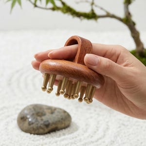 Może przedstawiać: Drewniane narzędzie do masażu z mosiężnymi końcówkami i uchwytem. Narzędzie trzymane jest w dłoni, z małym kamieniem i drzewkiem bonsai w tle. Narzędzie przeznaczone jest do masażu skóry głowy.