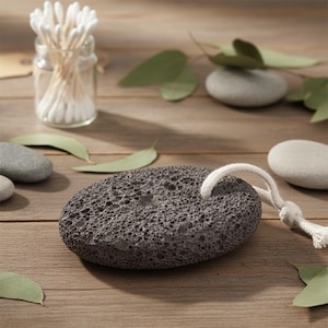 Puede incluir: Una piedra pómez gris oscuro con un lazo de cuerda blanca, rodeada de piedras grises lisas, hojas de eucalipto y un tarro de cristal con bastoncillos de algodón. La piedra pómez porosa es de forma ovalada.