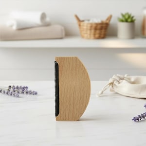 May include: A wooden lint brush with a curved handle and black bristles. The brush is made of light-coloured wood and is designed for removing lint and pet hair from clothing and fabrics. The brush is shown on a white surface.