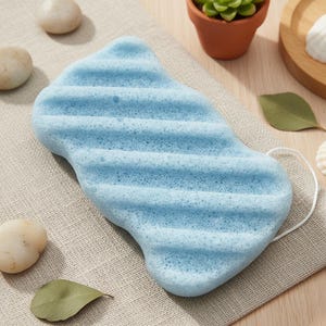 Pode incluir: Uma esponja de banho azul clara, texturizada, com ranhuras horizontais e uma argola para pendurar. Com formato de folha estilizada, repousa sobre um tecido de cor neutra. Pedras decorativas e uma pequena planta em vaso ao fundo.