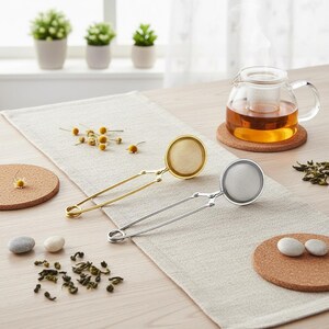 May include: Two tea infusers, one gold and one silver, with round mesh baskets and long handles, are displayed on a beige table runner. A glass teapot with tea steams in the background, along with tea leaves and chamomile flowers.