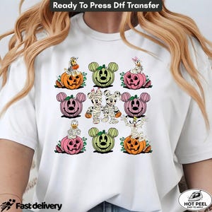 May include: A white t-shirt featuring a graphic print of various cartoon characters dressed as mummies and sitting on or next to colorful jack-o'-lanterns. The design includes orange, pink, and green pumpkins with character ears and faces.