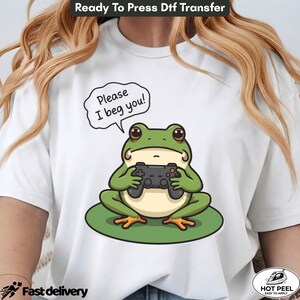 May include: A white t-shirt featuring a graphic of a green frog sitting on a lily pad while holding a black video game controller. Above the frog, a speech bubble contains the black text "Please I beg you!" in a simple, clear font.