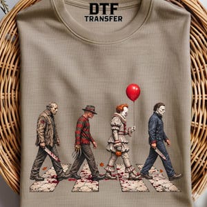 May include: A beige t-shirt featuring a graphic of four horror movie characters walking across a crosswalk. The characters include a masked figure with a blade, a striped sweater figure with clawed hands, a clown holding a red balloon, and a figure in a blue jumpsuit holding a knife. The text DTF TRANSFER is printed at the top.