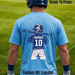May include: Light blue baseball jersey with a graphic of a baseball player holding a bat. The graphic includes the name "GUNNER" and the number "10". The text "Ready To Press" and "Custom Dtf Transfer" are also visible.