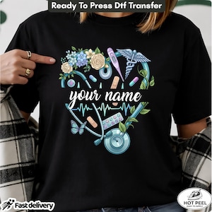 May include: A black t-shirt featuring a heart-shaped graphic composed of a stethoscope, medical instruments, flowers, and a butterfly. The text "your name" is printed in white script across the center of the heart design, surrounded by a blue heartbeat line.