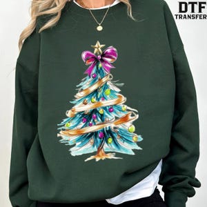 May include: A dark green crewneck sweatshirt featuring a graphic of a stylised Christmas tree. The tree is decorated with a large pink bow at the top, gold ribbon garlands, and small colourful ornaments in shades of green, pink, and blue.