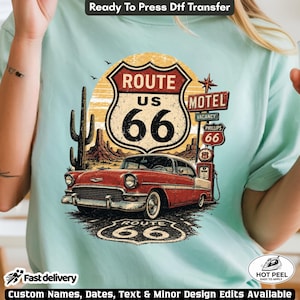 May include: A light mint green t-shirt featuring a graphic print of a red vintage car parked at a Route 66 gas station. The design includes a Route 66 shield sign, a motel sign, a cactus, and desert landscape elements with warm yellow tones.