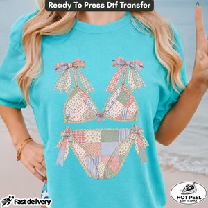 May include: A bright turquoise t-shirt featuring a graphic of a bikini set made from a patchwork of floral and gingham patterns in shades of pink, blue, and green. The bikini top and bottom are accented with bows at the straps and sides.