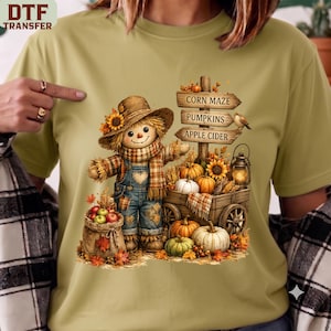 May include: A light olive green t-shirt featuring a graphic of a scarecrow wearing a plaid scarf and denim overalls. Beside the scarecrow is a wooden wagon filled with pumpkins, apples, and a lantern, with signs reading Corn Maze, Pumpkins, Apple Cider.