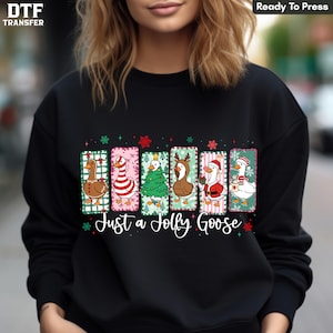 May include: A black long-sleeve sweatshirt featuring a graphic of six geese in individual rectangular panels. The geese are dressed in holiday themes including a gingerbread pattern, candy cane stripes, a Christmas tree, reindeer antlers, a Santa suit, and a scarf. The text reads Just a Jolly Goose.