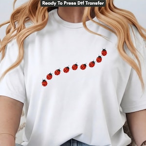 May include: A white short-sleeved t-shirt featuring a diagonal graphic print of nine red ladybugs with black spots. The ladybugs are arranged in a curved line across the chest area of the shirt, moving from the lower left toward the upper right.