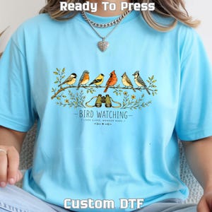 May include: A light blue t-shirt featuring a graphic of six colorful birds perched on a branch above a pair of binoculars. The text reads BIRD WATCHING LOOK CLOSE, WONDER MORE. The shirt has a crew neckline and short sleeves, with a heart-shaped pendant.
