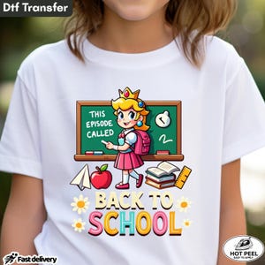 May include: A white t-shirt featuring a graphic of a cartoon princess with a crown and pink outfit standing in front of a green chalkboard. The board reads THIS EPISODE CALLED. Below are an apple, books, and the text BACK TO SCHOOL in colorful letters.