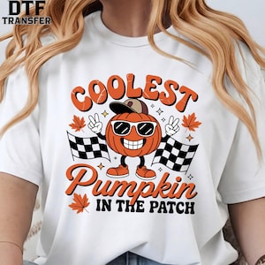 May include: A white t-shirt featuring a graphic of an orange pumpkin wearing sunglasses and a baseball cap. The pumpkin is making peace signs with its hands, flanked by two checkered racing flags and orange maple leaves. Text reads COOLEST PUMPKIN IN THE PATCH.