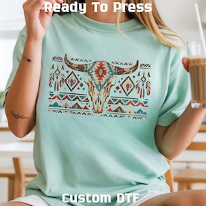 May include: A light mint green t-shirt featuring a graphic of a bull skull with geometric patterns in red, blue, and tan. The text "Ready To Press" appears at the top and "Custom DTF" at the bottom. The design has a distressed, weathered texture.