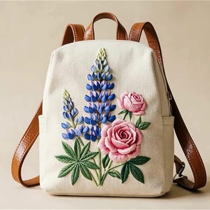 May include: A cream-coloured canvas backpack featuring an embroidered design of blue lupine flowers and pink roses with green leaves. The bag includes brown leather straps and a top handle, with the IJD Leather logo visible in the top left corner.