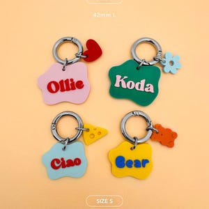 May include: Four colorful keychains with silver rings. Each keychain has a unique shape and color, with names like "Ollie," "Koda," "Ciao," and "Bear." The keychains also feature small charms like a heart, flower, cheese, and a gummy bear.