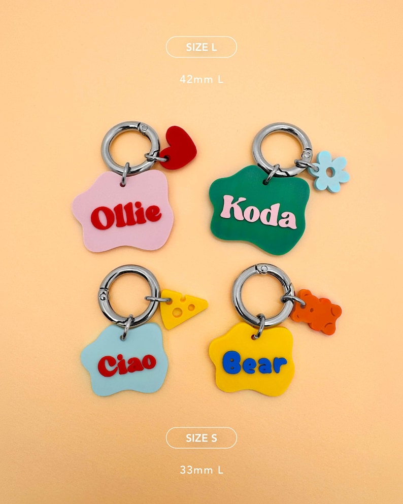 May include: Four colorful keychains with silver rings. Each keychain has a unique shape and color, with names like "Ollie," "Koda," "Ciao," and "Bear." The keychains also feature small charms like a heart, flower, cheese, and a gummy bear.