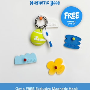 May include: A collection of colorful magnetic hooks and pet tags. The main hook features a tennis ball tag with the name "Luna" and a miniature tennis racket. Other hooks are shaped like a flower, cloud, and abstract shapes. The text "Magnetic Hook" and "FREE" are also visible.