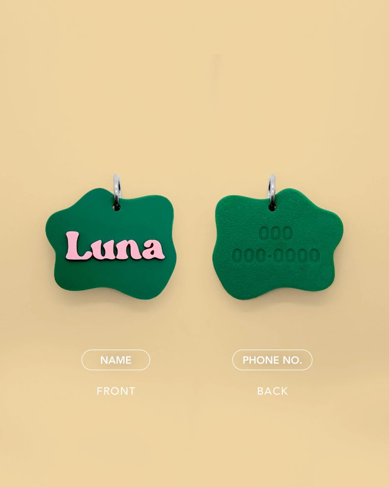 May include: Two green pet tags with silver hoops. The front tag has the name "Luna" in pink letters. The back tag has a phone number embossed. The tags are a unique, irregular shape. The text "NAME" and "PHONE NO." are below the tags.