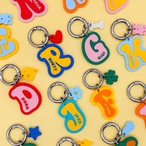 May include: A collection of colorful, personalized keychains with names like "Nova," "Archie," and "Bella." Each keychain features a unique shape, vibrant colors, and a silver keyring. Some keychains have small charms like hearts and stars.