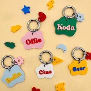 May include: A collection of colorful keychains with silver rings. Each keychain features a unique shape and name: Ollie, Koda, Bella, Ciao, and Bear. Additional decorative shapes include stars, hearts, and flowers.
