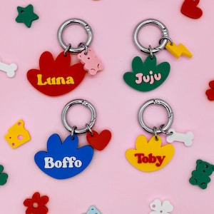 May include: Four keyrings with colourful charms, including a red heart, a yellow lightning bolt, and a white bone. Each keyring has a different name: Luna, Juju, Boffo, and Toby. The background is pink.