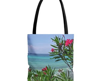 May include: Tote bag with a beach scene: turquoise sea, blue sky, and pink flowers. The bag has a black handle. The background shows a white fence. Ideal for carrying items, this bag is a stylish accessory.