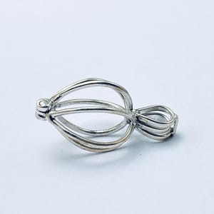 May include: A silver-colored pendant with a cage-like design. The pendant features a series of curved, parallel bars that form an open, oval shape. The design includes a loop at one end for a chain.