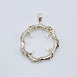 May include: A silver pendant with a circular design resembling intertwined twigs. The pendant has a loop at the top for a chain and four small prongs inside the circle. The pendant is set against a white background.