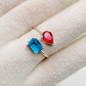 Puede incluir: Un anillo abierto de plata con dos piedras preciosas distintas. Una piedra es de color azul vibrante con corte rectangular, mientras que la otra es una piedra roja con forma de pera. La banda está hecha de metal plateado pulido y brillante.