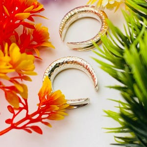 Könnte beinhalten: Zwei silberne, halbmondförmige Ohrringe mit gehämmerten Texturen. Die Ohrringe werden auf einer weißen Oberfläche präsentiert, umgeben von bunten Kunstblumen in Orange, Gelb, Lila und Grün. Die Ohrringe sind ein Schmuckstück.