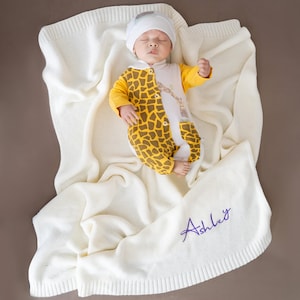 May include: A sleeping baby wearing a yellow and brown giraffe-patterned onesie and a white hat, resting on a cream-colored knitted blanket. The blanket is personalized with the name "Ashley" in purple script.