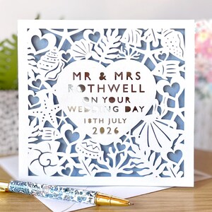 May include: A white wedding card with a laser-cut design featuring seashells, hearts, and marine elements. The card reads "MR & MRS ROTHWELL ON YOUR WEDDING DAY 18TH JULY 2026". A pen with a floral pattern lies on the envelope, next to the card.
