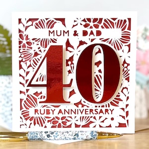 May include: A square white card with a red floral design and the words "MUM & DAD" at the top. The number "40" is in the center, with "RUBY ANNIVERSARY" at the bottom. A pen with a floral design rests in front of the card.