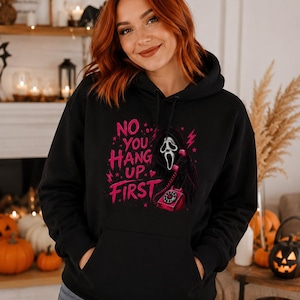 May include: A black hooded sweatshirt featuring a graphic print of a masked figure holding a pink rotary telephone. The text reads NO YOU HANG UP FIRST in pink lettering, accompanied by pink lightning bolts and star accents on the front chest.