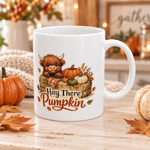 May include: A white ceramic mug featuring a graphic of a fluffy brown Highland cow sitting on a hay bale with orange and white pumpkins. The text "Hay There Pumpkin" is printed in brown script and block lettering below the illustration.