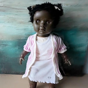 Peut inclure: Poupée vintage à la peau foncée, aux cheveux noirs bouclés et vêtue d'une robe blanche. La poupée porte un cardigan rayé rose et blanc. Le visage de la poupée a une expression solennelle. La poupée se tient devant un fond texturé.